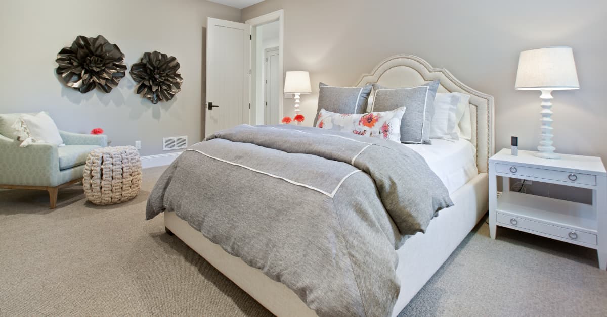 Cama matrimonial con cabecero tapizado en tonos beige, edredón gris y cojines decorativos, en un dormitorio moderno con lámparas de mesa y paredes neutras.